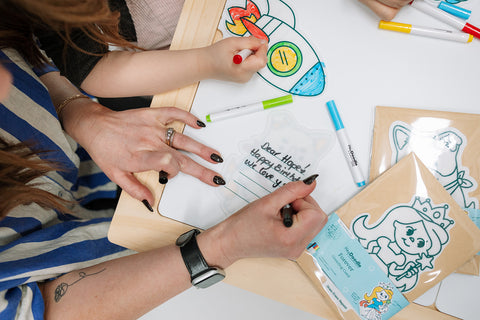 Two kids use colorful markers on silicone mats—one colors a rocket, the other writes a note. Mermaid-themed mats and marker sets nearby.