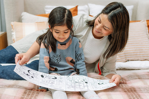 A woman and young girl smile on a bed, enjoying a colorful silicone activity mat with playful cartoon designs in gentle lighting.