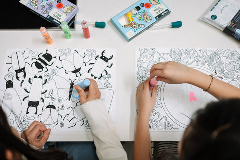 Two people color insect-themed silicone mats with paint markers. Colorful markers and art boxes are scattered on the white tabletop.