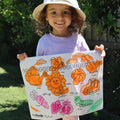 Little girl wearing hat holding mat that is colored in orange, pink and green, mat features garden elements like sunflower, watering can, butterfly, vegetables and fruits, shovel