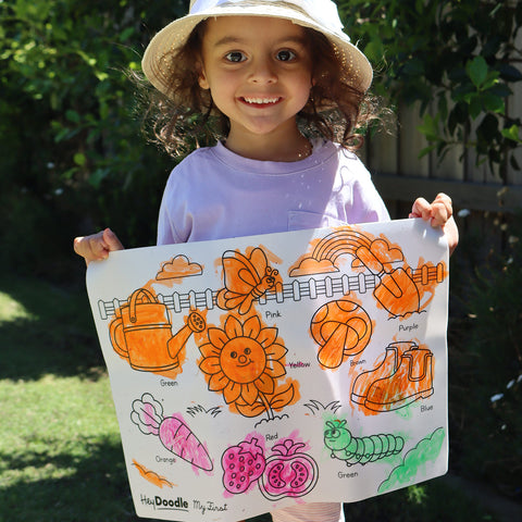 Little girl wearing hat holding mat that is colored in orange, pink and green, mat features garden elements like sunflower, watering can, butterfly, vegetables and fruits, shovel