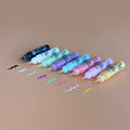 Eight HeyDoodle Glitter Peel & Stick Paint bottles on a tan surface, each with a wavy line of vibrant silicone color in front.