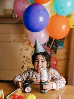 A smiling young child in a party hat colors on a silicone activity mat at a table, with snack containers, balloons, and confetti nearby.