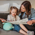 A woman and young girl smile on a bed, coloring a HeyDoodle silicone reusable mat as toys and a bear add to the playful vibe.
