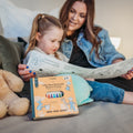 A woman and girl sit on a bed coloring a HeyDoodle silicone mat, with a teddy bear, colorful crayons, and reusable designs nearby.