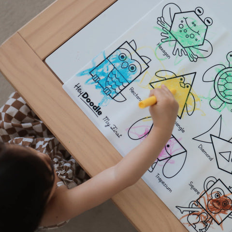 Kid coloring with yellow crayon on mat that shows geomatrical shaped animals like frog, owl, monkey, bird