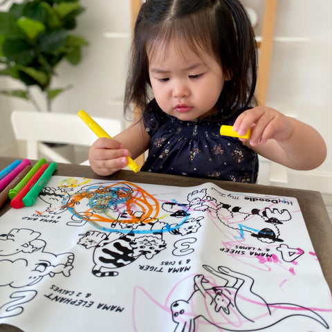 Little girl holding yellow crayon, coloring on mat that shows mama animals and the little baby animals