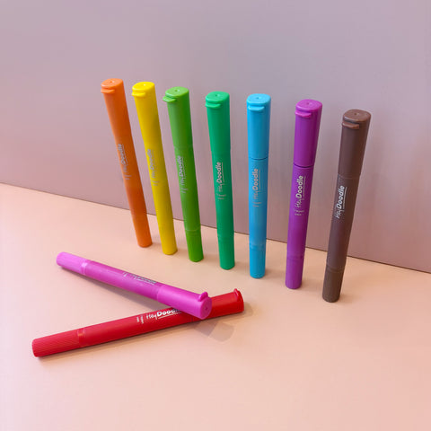 Eight HeyDoodle silicone crayon pens—yellow, orange, green, light green, teal, blue, purple, brown—stand in a row; red and pink lie in front on a light pink surface.